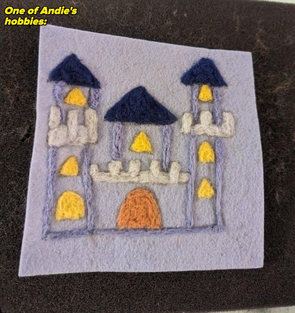 A felt art piece depicting a simple, abstract castle with two towers and a main entrance. The design includes basic geometric shapes and minimal detail