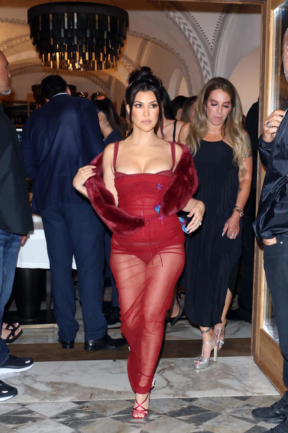 Kourtney in a burgundy-red sheer long dress revealing a red bodysuit underneath. The sheer dress has sporadic blue floral applique. She has. burgundy fur stole around her arms and red criss-cross strappy heels.