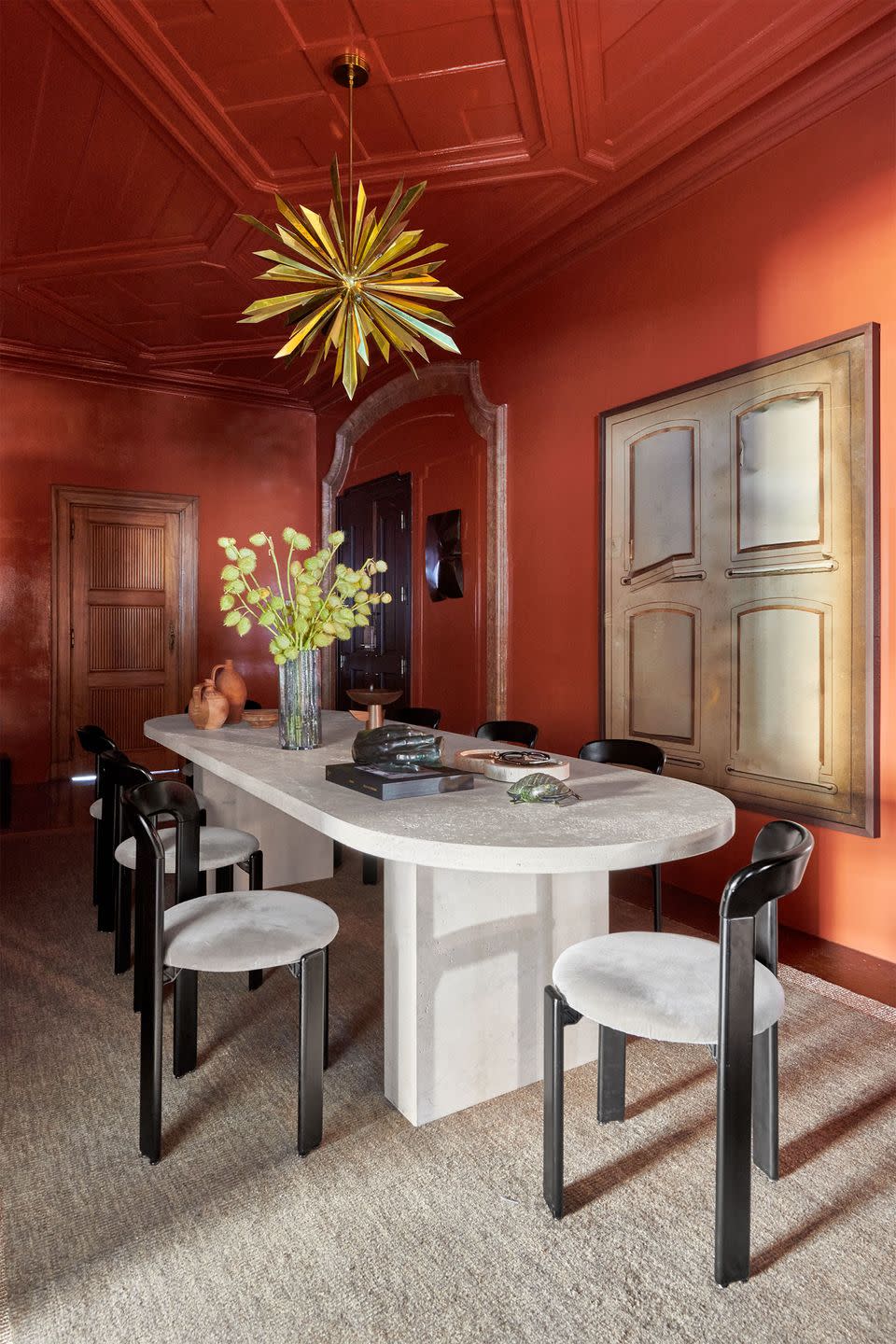 in a dining room is a long white travertine table with rounded ends, eight minimalist black chairs with round seats, rust colored walls, artwork resembling windows with shades, and a spiky gold light pendant