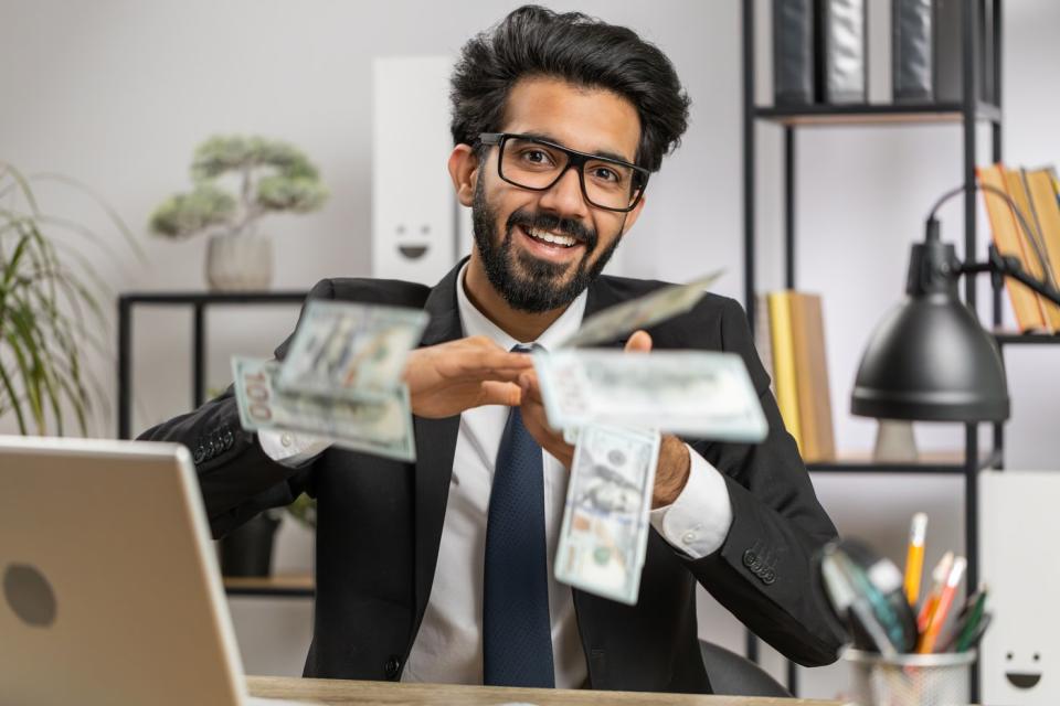 A person in a business suit seated at a desk smiles while throwing $100 bills. 