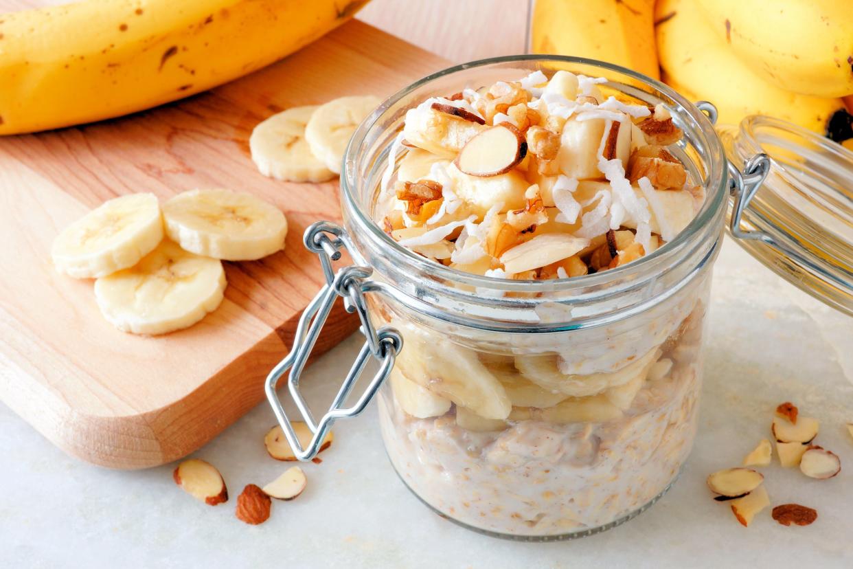 Overnight oats in a snap lid glass jar on the left, cut banana on a cutting board on the left, surrounded by bananas on a white marble table