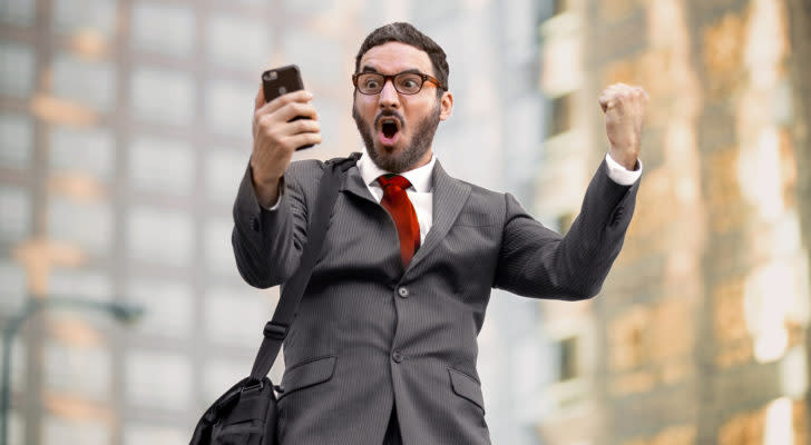 A photograph of an excited man in a suit looking at a cellphone in one hand while raising his other hand in a cheering motion.
