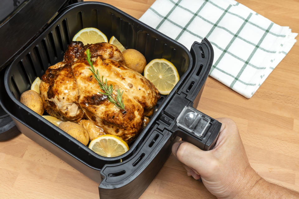 A person's hand lifting the lid of an air fryer containing a roasted chicken and potatoes
