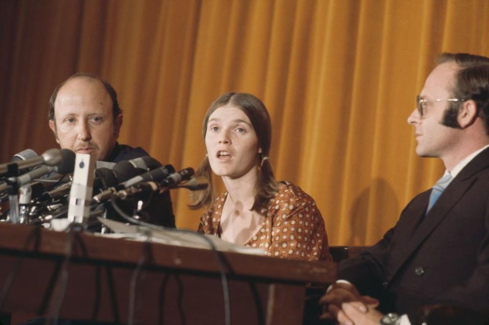 a woman in a brown dotted dress speaks into several microphones at a table, seated next to two men in suits