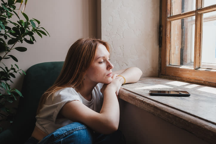 Woman sitting by a window, resting chin on her hand, gazing outside, phone on the windowsill