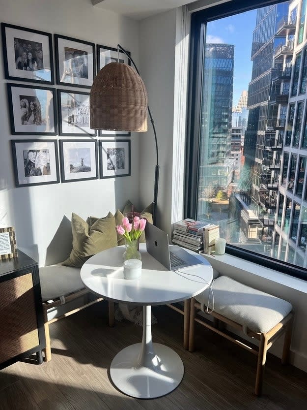 A cozy home office with a small round table, a bench, floor lamp, and a view of city buildings through a large window