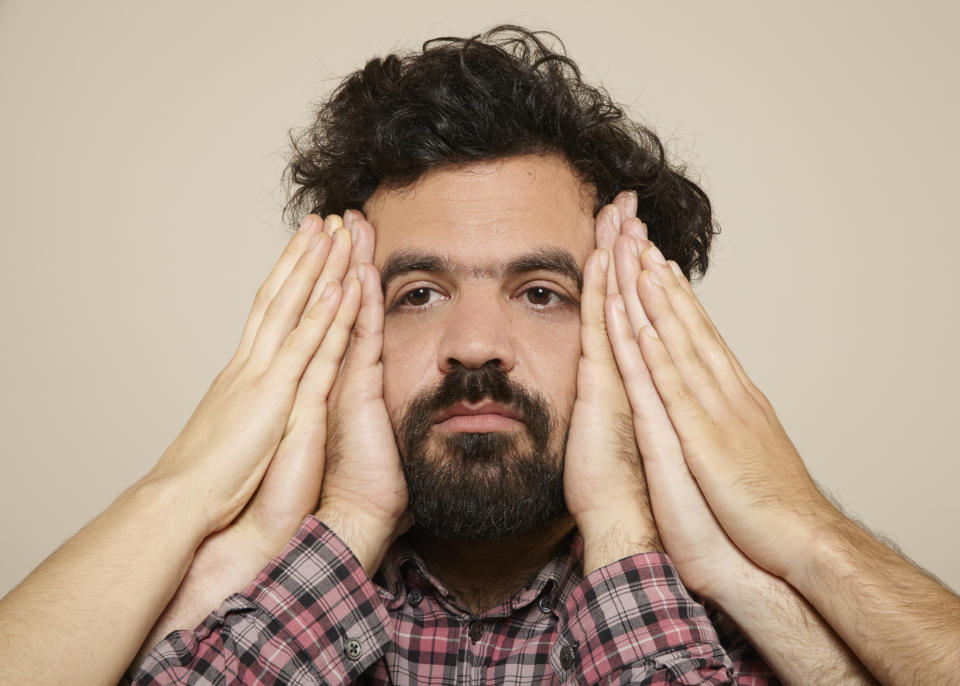 Bearded man with several pairs of hands, including his own, pressed on either side of his face