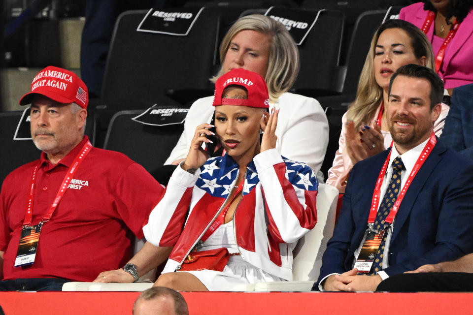 Mehrere Personen, darunter Mel B, sitzen bei einer Veranstaltung. Mel B trägt ein patriotisches Outfit und eine rote Trump-Mütze, während sie telefoniert