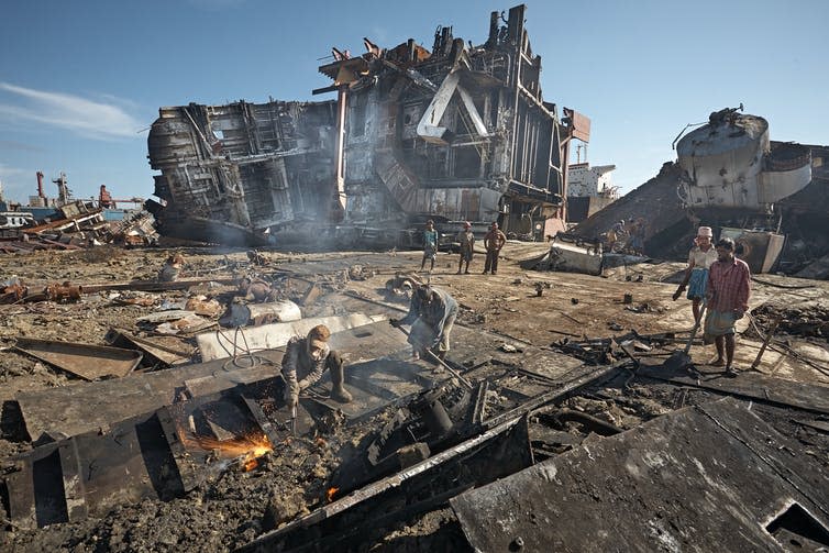 People dismantling parts of a ship, large ship on its side in the background.