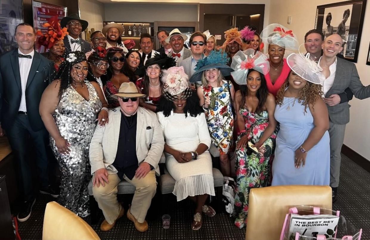 Several dozen people wearing flamboyant outfits with hats pose together in a room.