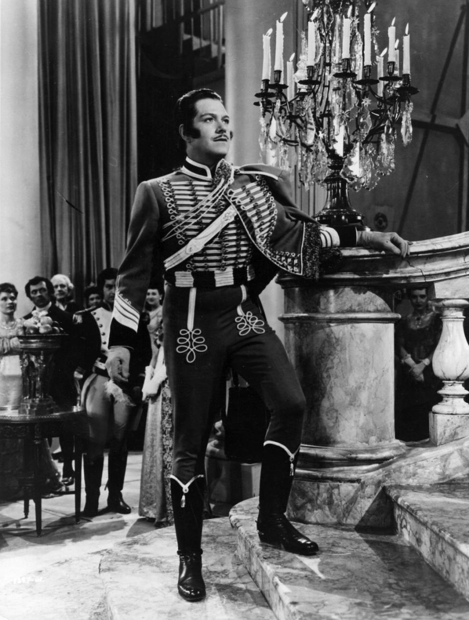 a black and white photo of nelson eddy on a movie set, wearing an aristocratic period costume, looking up from the bottom of a staircase, with several extras standing behind him