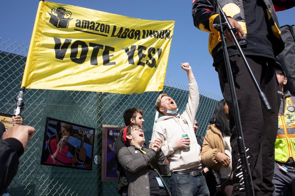 Amazon warehouse workers cheer outside a fence and hold a picket sign that reads: Amazon Labor Workers Vote Yes.