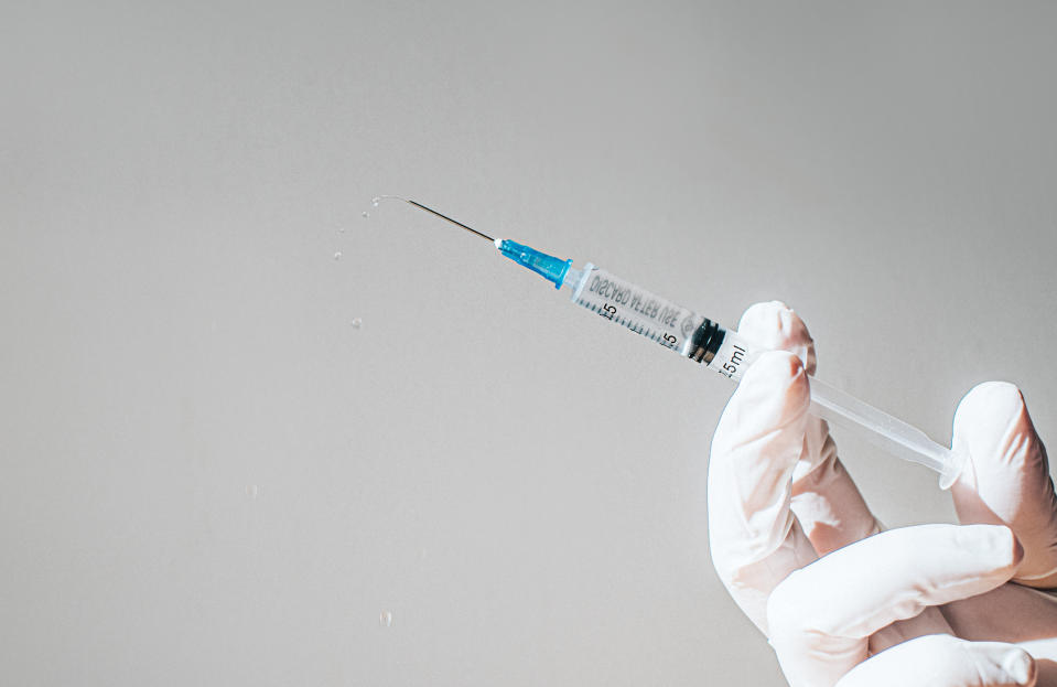 A gloved hand holding a filled syringe with a drop of liquid at the tip. The background is plain and neutral
