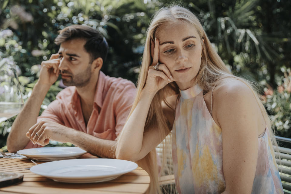 A woman and a man sit at a table outdoors, appearing distant and thoughtful, with empty plates in front of them