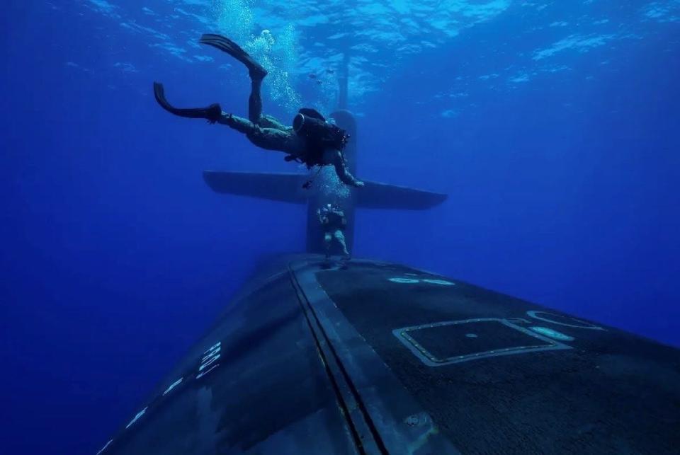 Two US Marines dive in blue water above a US submarine in the open ocean.