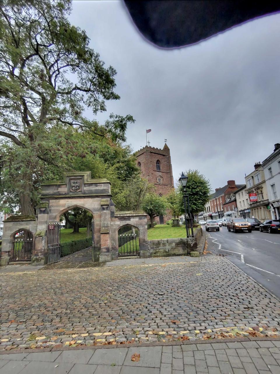 A church on a highstreet on a cloudy day, a flag flies above it and cars are driving past