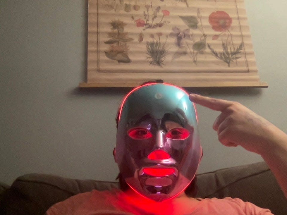 A person pointing to a sticker-demarcated button on an LED face mask against a blue wall with a flower poster on it.