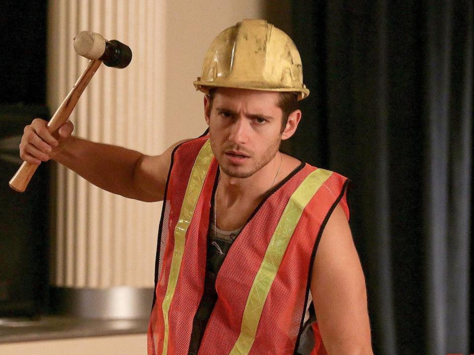 Julian Morris in an orange construction best and yellow hard hat holding a mallet leaning on a plank