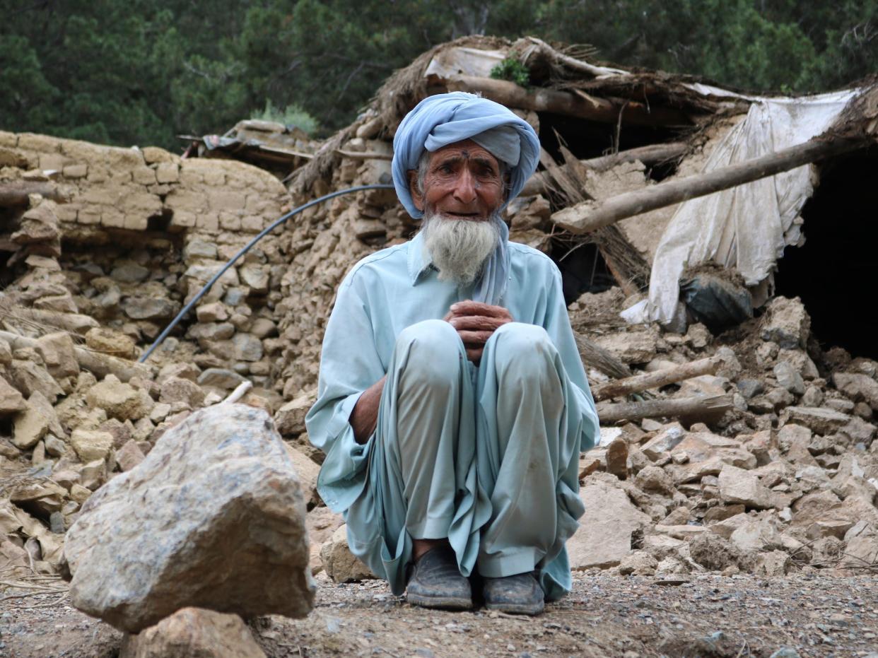 man in blue with white beard sits amid rubble from earthquake crumbled home