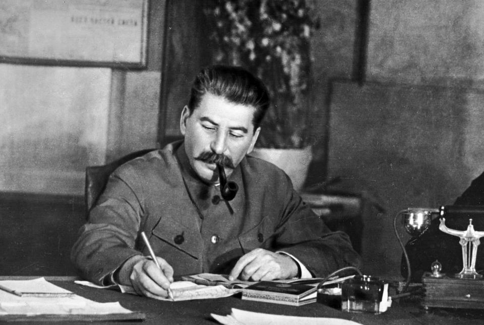 A black and white photo of Joseph Stalin writing on a piece of paper and smoking a pipe at his desk.