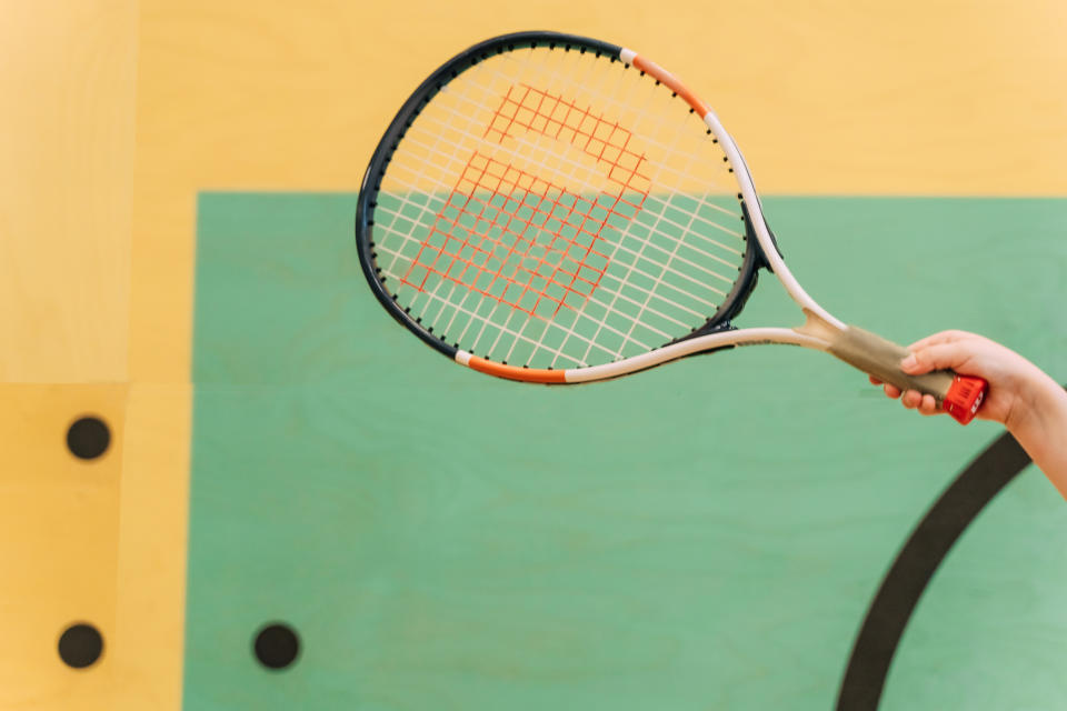 A close-up of a person's hand holding a tennis racket against a light background, including part of a tennis court's green area