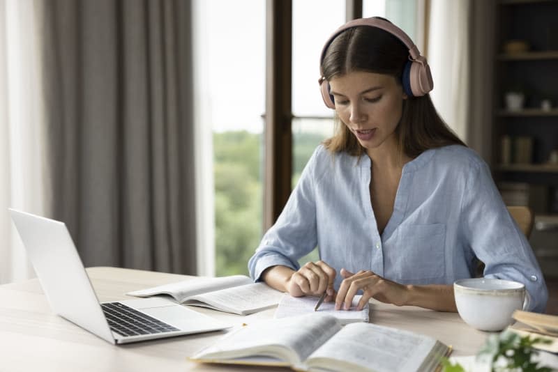 Focused engaged student girl in big headphones studying foreign language, listening audio lesson at laptop, reading notes out loud, doing exercises from open book, school, college homework task