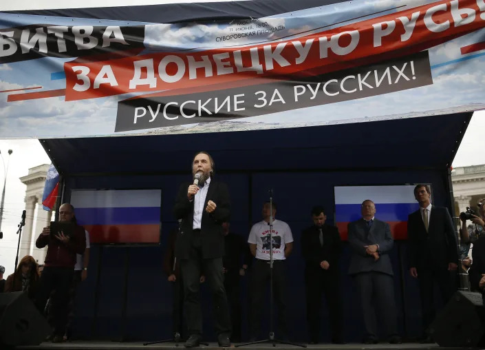 Alexander Dugin speaks into a microphone while standing on a stage below a Russian language banner.