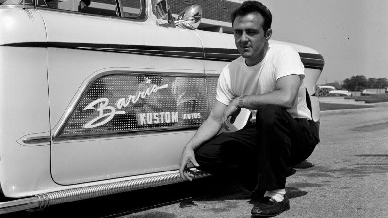 A black and white photo of George Barris kneeling next to a pickup truck