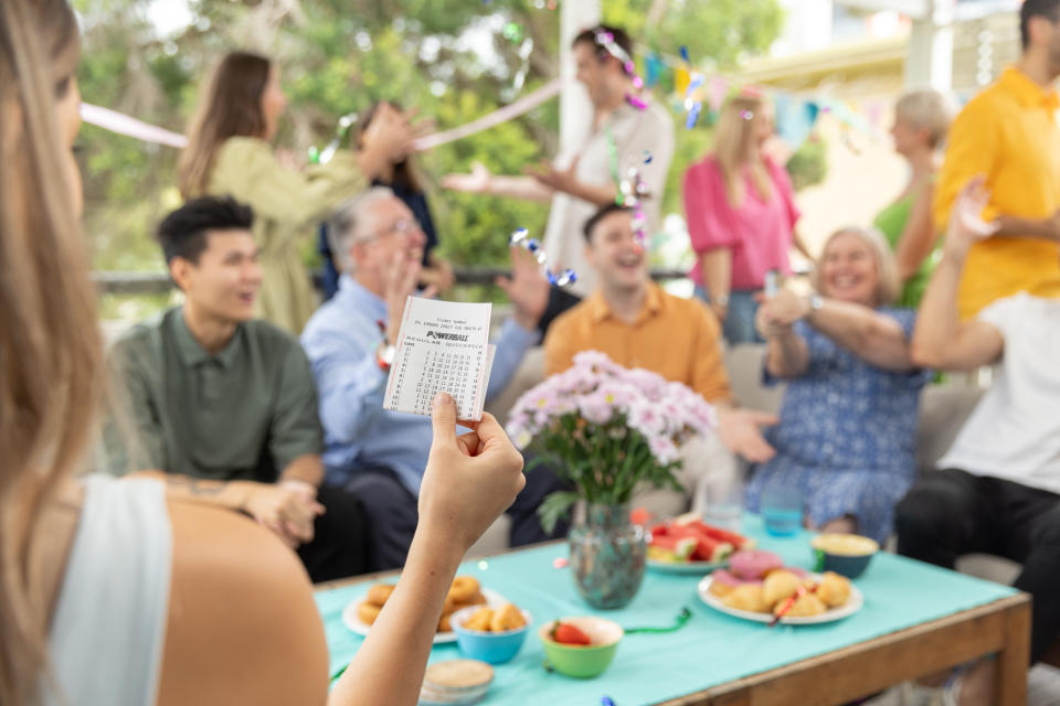 Group of people celebrating a Powerball win as woman holds ticket. 