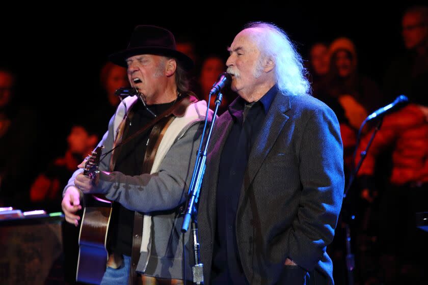 Two men, one a jacket holding a guitar and another in a suit, singing on a stage