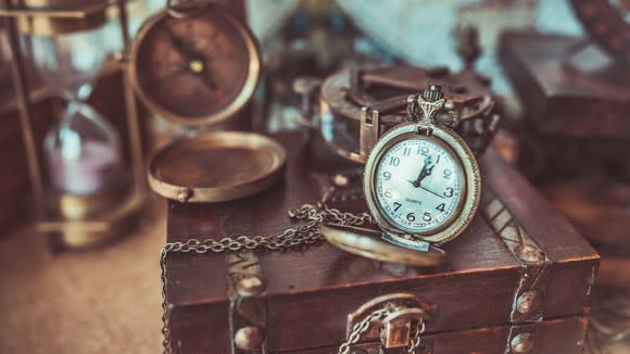 A number of antiques, including an hour glass, a compass, and a pocket watch set atop an old wooden box.