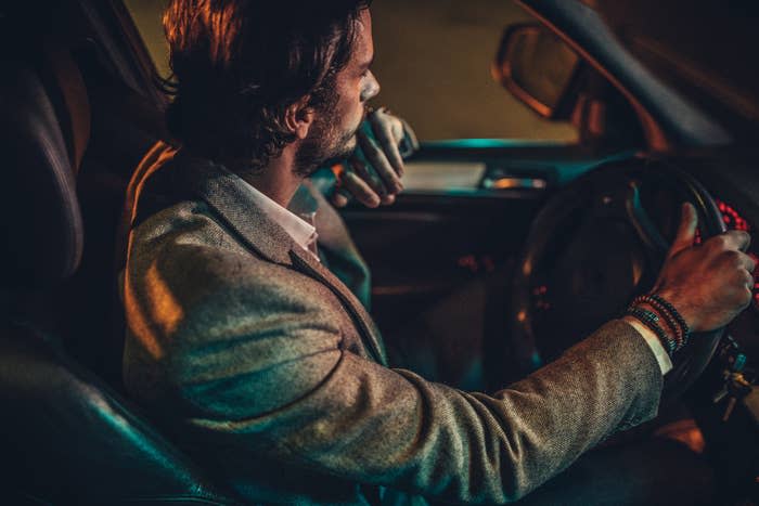 Man in a blazer driving a car at night, looking to the side, hand on steering wheel