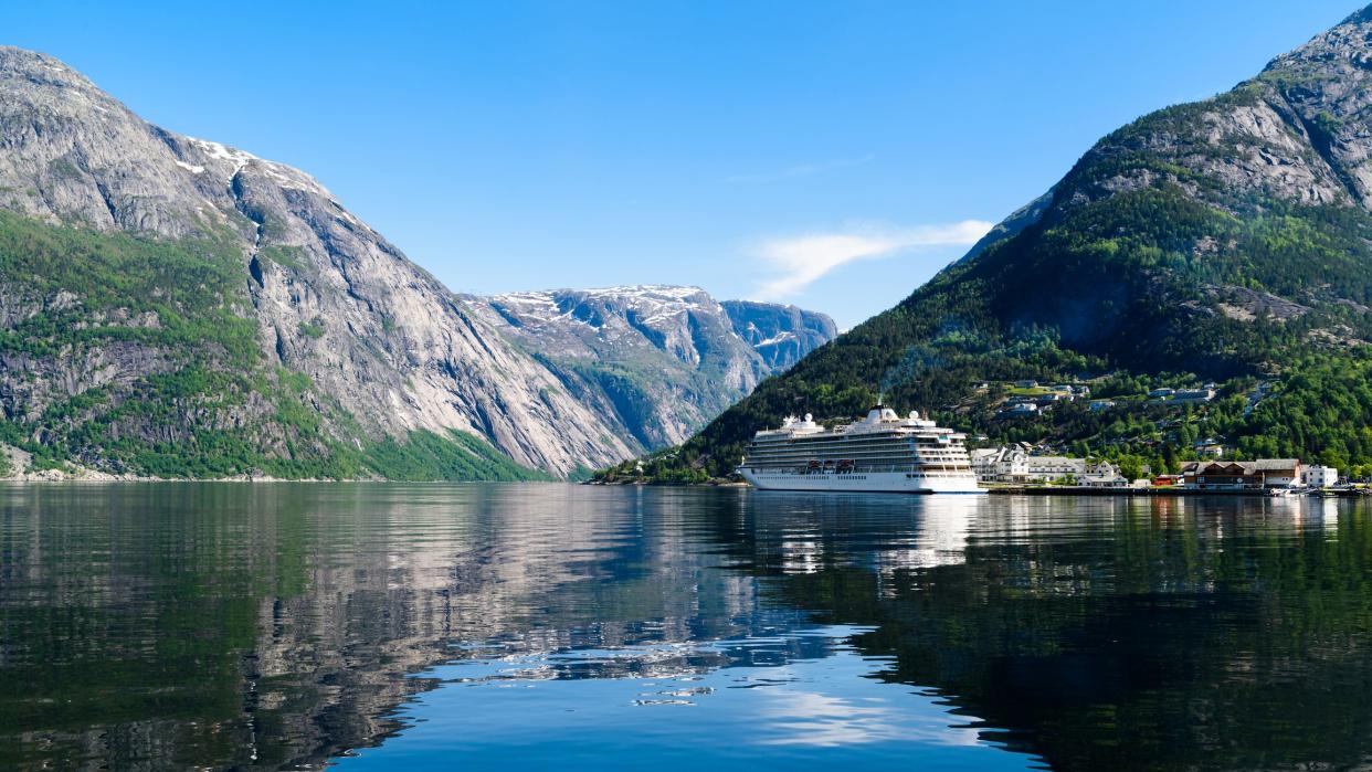 The village Eidfjord in Hordaland, Norway on a very tranquil and sunny day. A cruise ship is moored outside the little town in the fjord and the surrounding mountains reach for the blue sky.