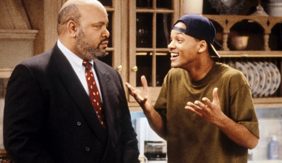 James Avery, in a suit, looks serious while Will Smith, in a t-shirt and backwards cap, appears animated and expressive in a kitchen setting from "The Fresh Prince of Bel-Air."