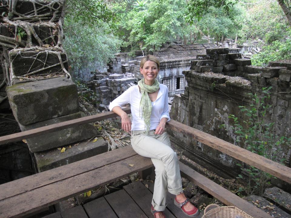 A woman sits on a wooden bench in front of a scene e of ruins in a forest in Cambodia