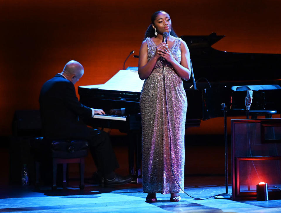 Vocalist Samara Joy performs onstage in a floor-length glittery dress. A piano and pianist are in the background.