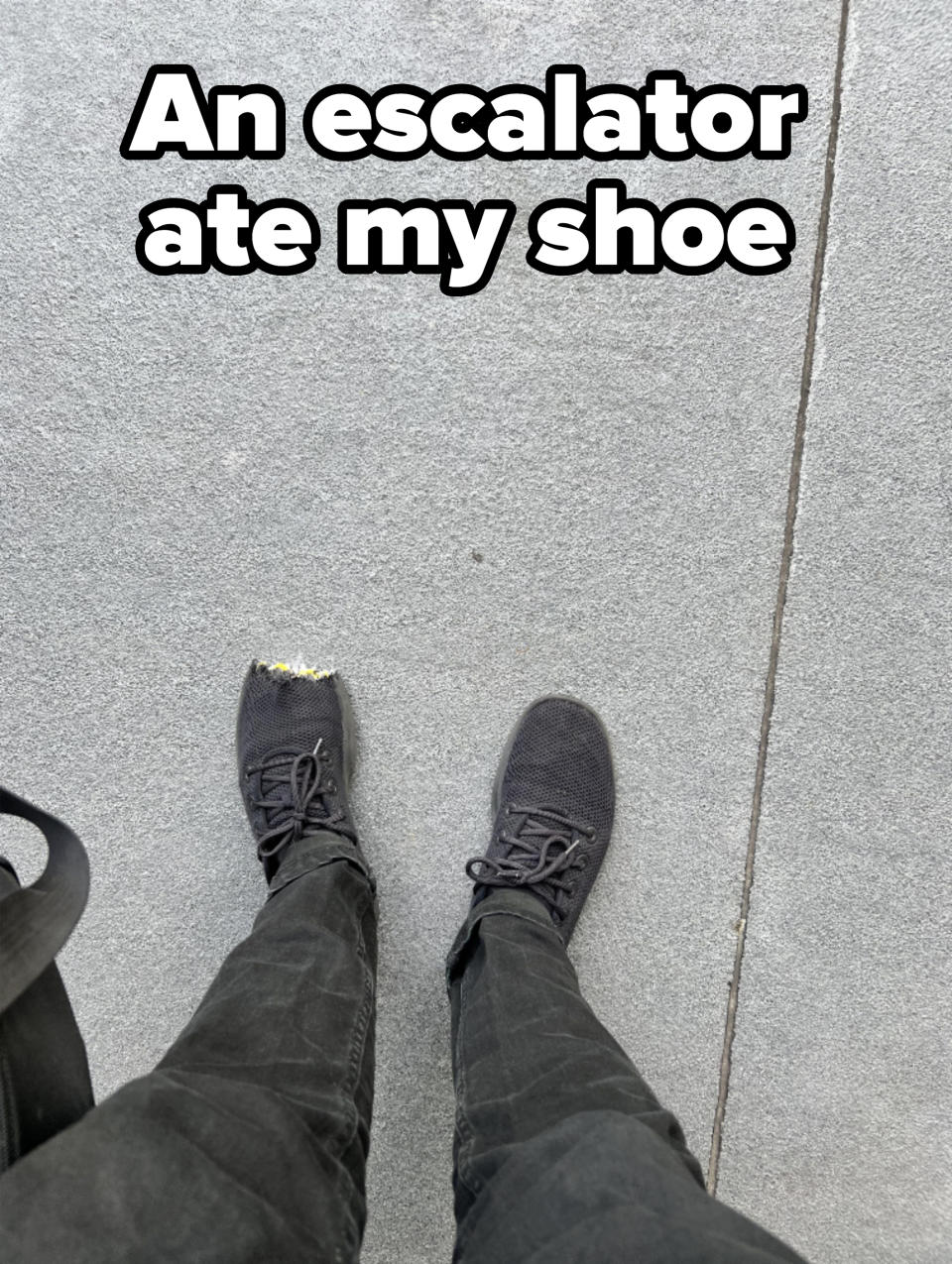 Person wearing black sneakers with one sneaker's toe area visibly worn out, standing on a concrete surface. Only their legs and feet are visible