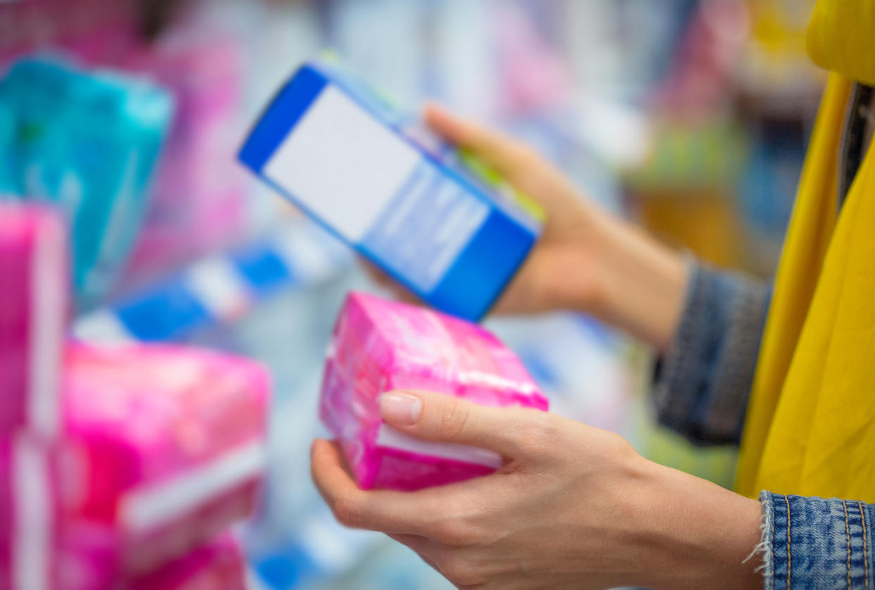 A woman holds two different packages of menstrual hygiene products in her hands in a store aisle. 