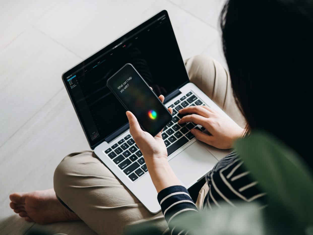 Young Asian woman using AI assistant on smartphone while using laptop working from home in the living room