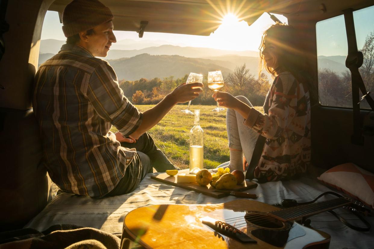 Sitting in the back of their camper van with the door open, a young couple are enjoying a picnic and a glass of wine, with the sun setting in front of them