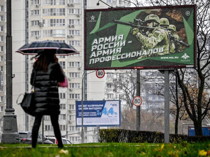 A pedestrian under an umbrella walks under a poster showing three Russian soldiers, with the slogan reading 'Army of Russia, army of professionals"