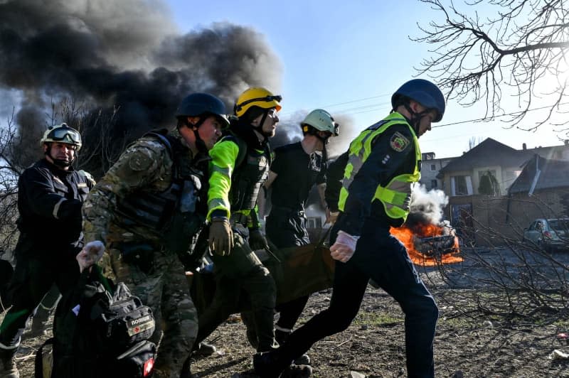 Rescue workers and police carry an injured man on a stretcher while a column of smoke rises in the background. -/Ukrinform/dpa