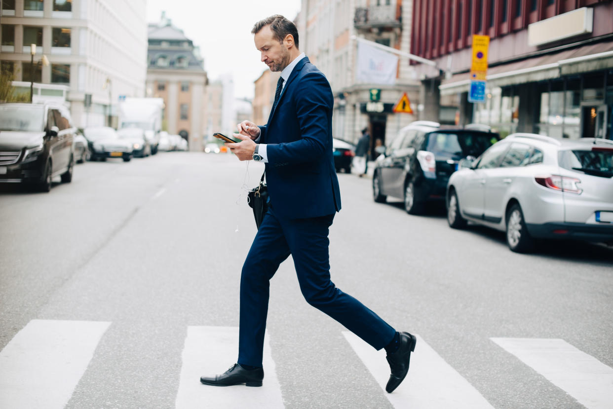 Full length side view of mature businessman crossing street while using smart phone in city