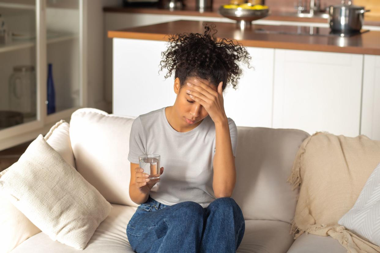 Unhappy African American woman sitting on the couch with a glass of water feeling bad, experiencing headache and dizziness due to illness