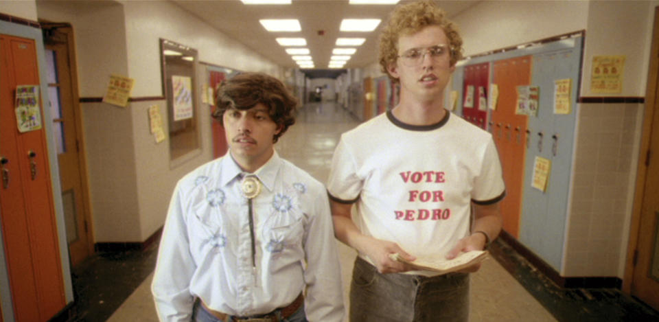 Two characters from the film 'Napoleon Dynamite' stand in a school hallway; one wears a 'Vote for Pedro' shirt