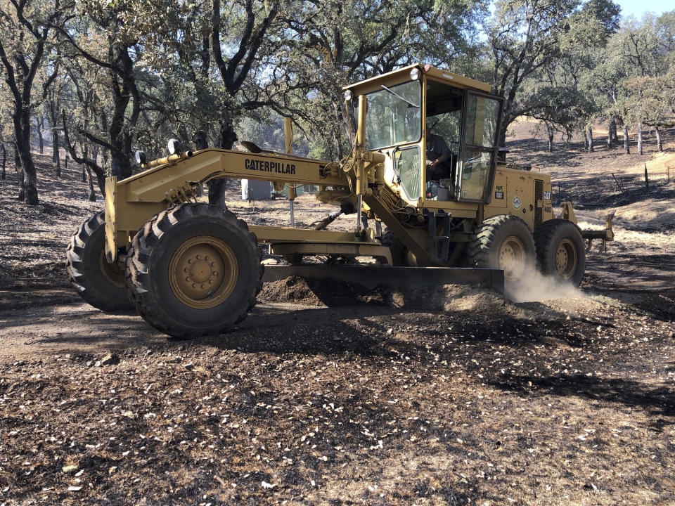 In this Friday, Aug. 10, 2018 photo, Jack Hattendorf steers a road grader to repair a dirt path near Lakeport, Calif. Even as flames continue chewing through forestland nearby, Hattendorf and others are working to repair the damage wrought not by flames but by firefighters trying to stop them. (AP Photo/Jonathan J. Cooper)