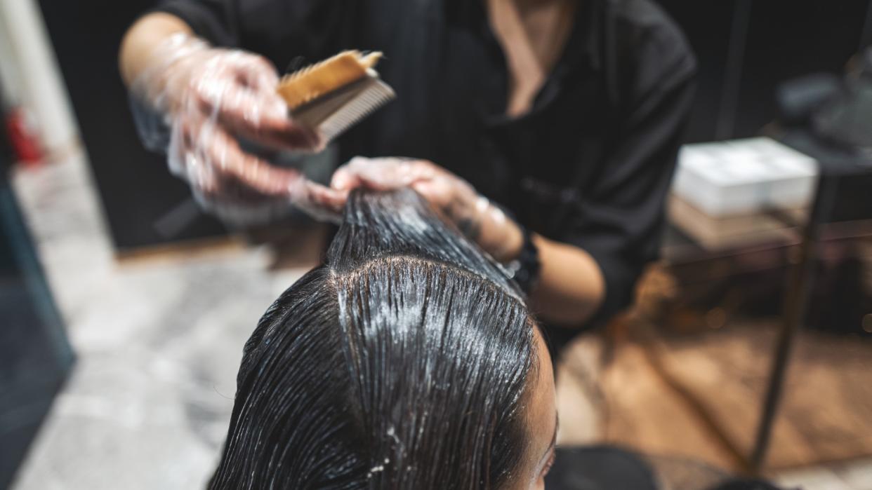  Close-up image of a hairdresser combing a white cream through a woman's long black hair in a salon. The woman's head in the foreground is in focus but the rest of the image is slightly blurred. 