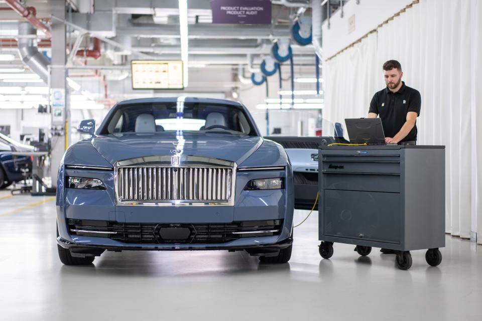 a man on a computer works next to blue car in factory