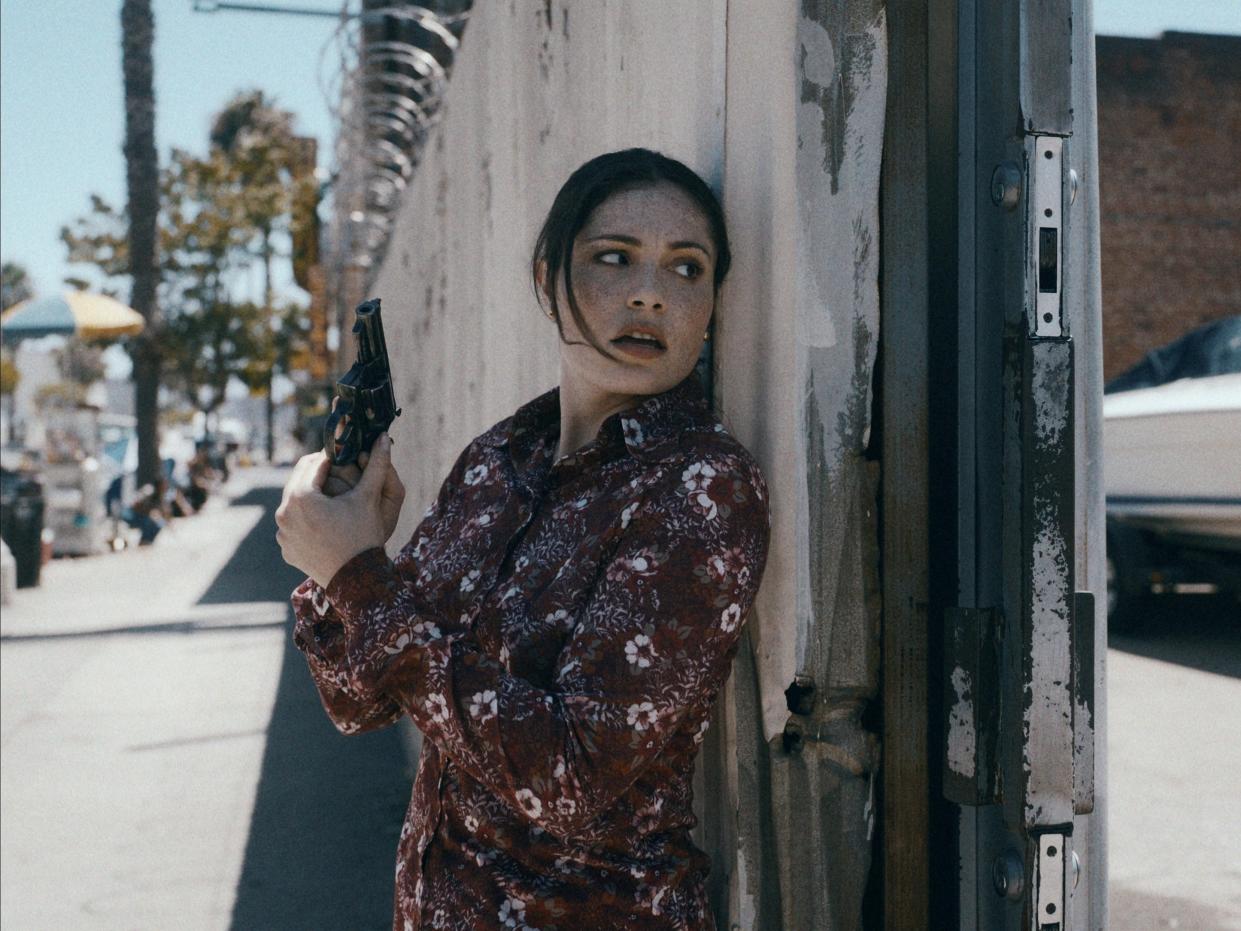 juliana aidén martinez as june hawkins in griselda. she's wearing a floral shirt and standing with ehr back against a wall, holding a gun