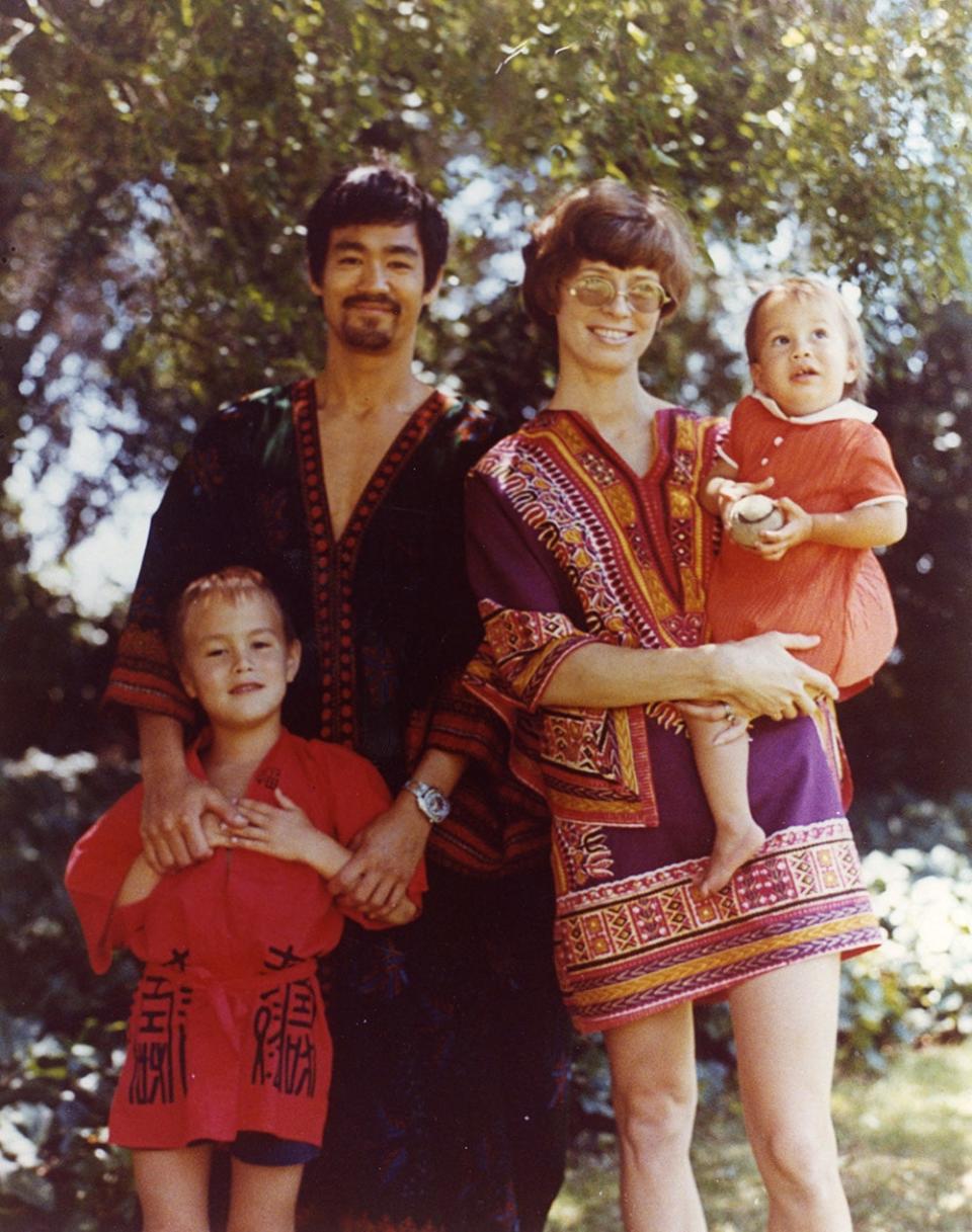 Family portrait, Bruce, Brandon, Linda, Shannon. Bruce is wearing a dark kaftan, beard, smiling, hand on Brandon, while Linda holds Shannon. Tree and grass in background.